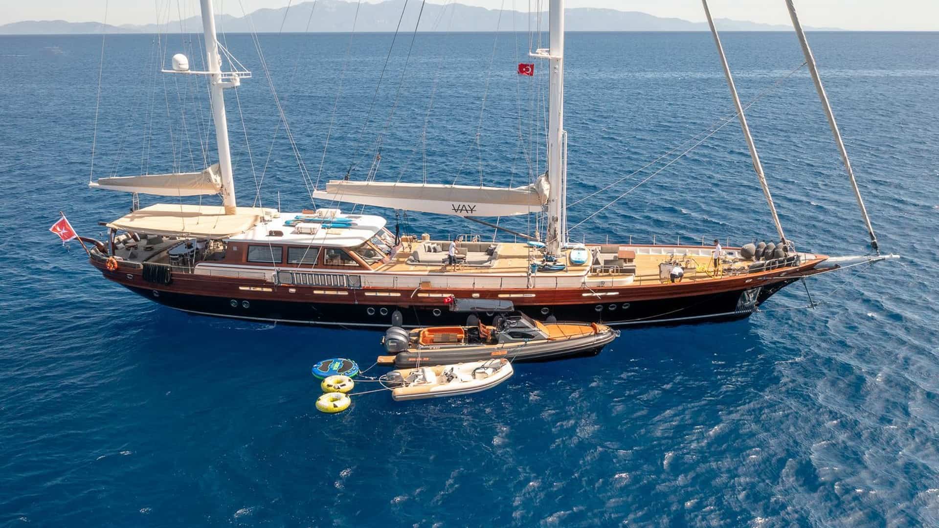 A large wooden sailing yacht anchored in blue ocean with a smaller inflatable dinghy tied nearby and people on deck.