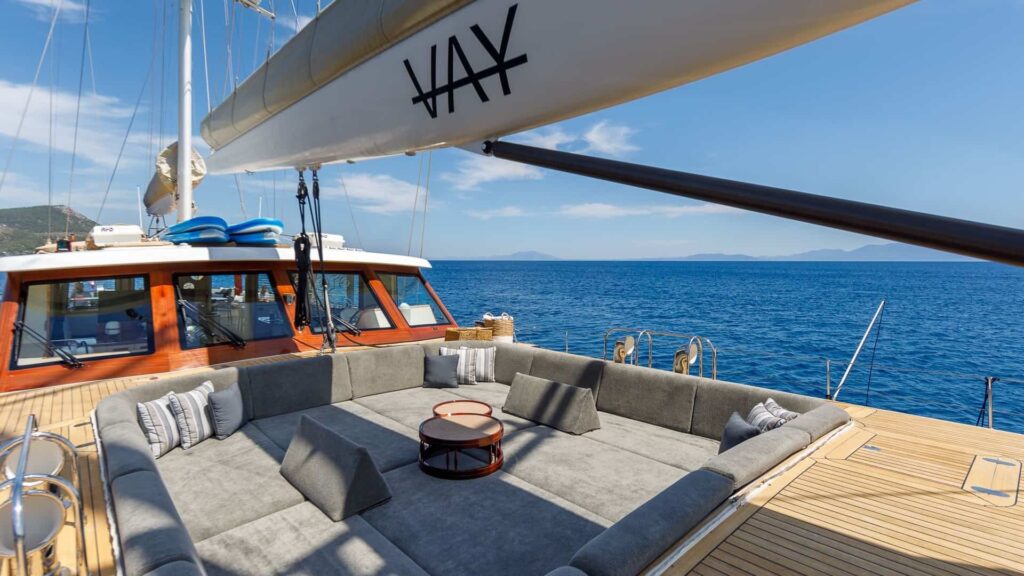Luxurious yacht deck with grey cushioned seating and round tables, overlooking blue ocean and distant hills.