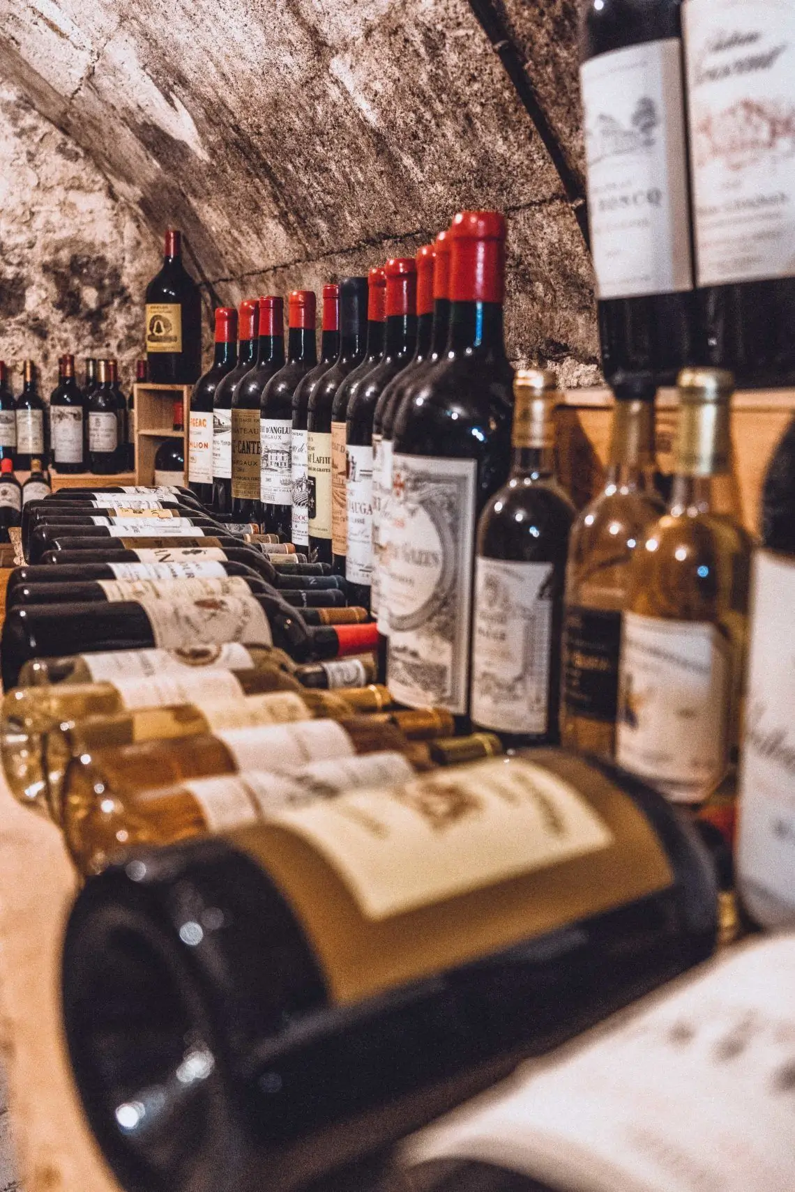 Row of wine bottles lined up on shelves against a rustic stone wall in a cellar. Red-capped bottles dominate the row, with blurred foreground bottles nearby.