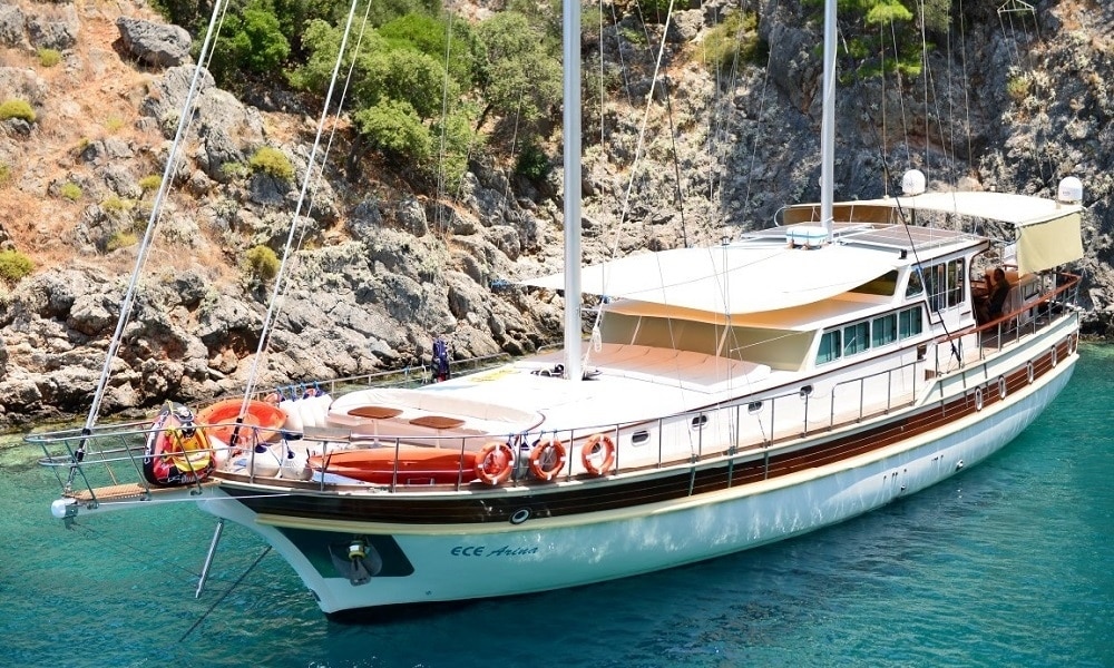 Luxurious white sailing yacht anchored near a rocky, pine-covered coastline with turquoise water and lifebuoys on deck