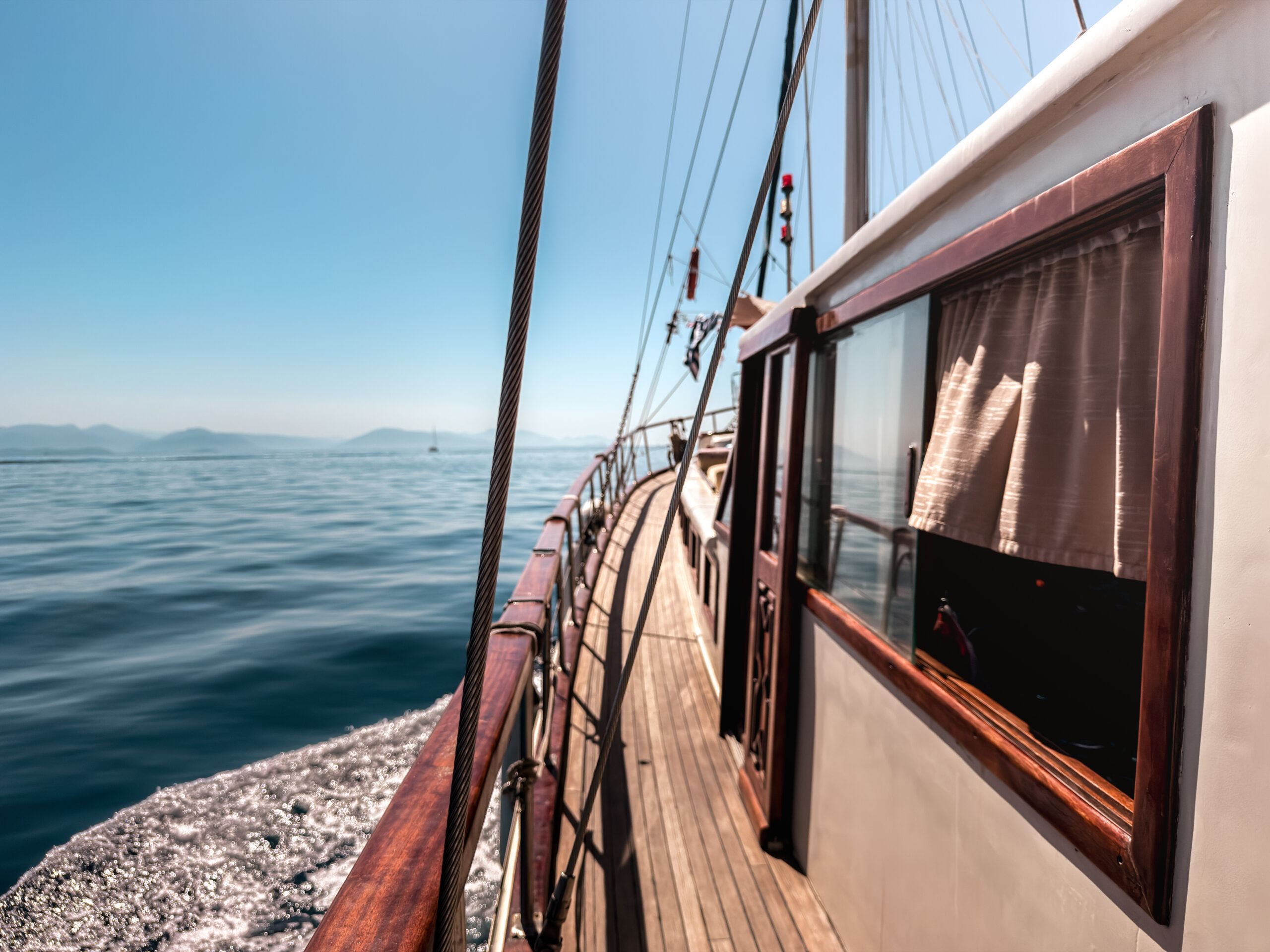 Deck of a wooden sailing boat with rigging and ropes, calm blue sea and distant mountains on the horizon