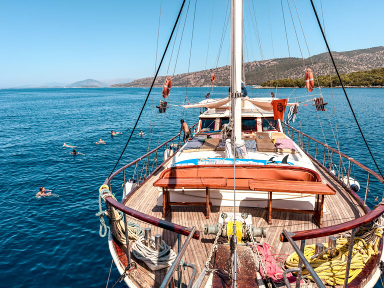 View from a sailing yacht deck watching swimmers in bright blue sea with a rocky coastal landscape in the distance.