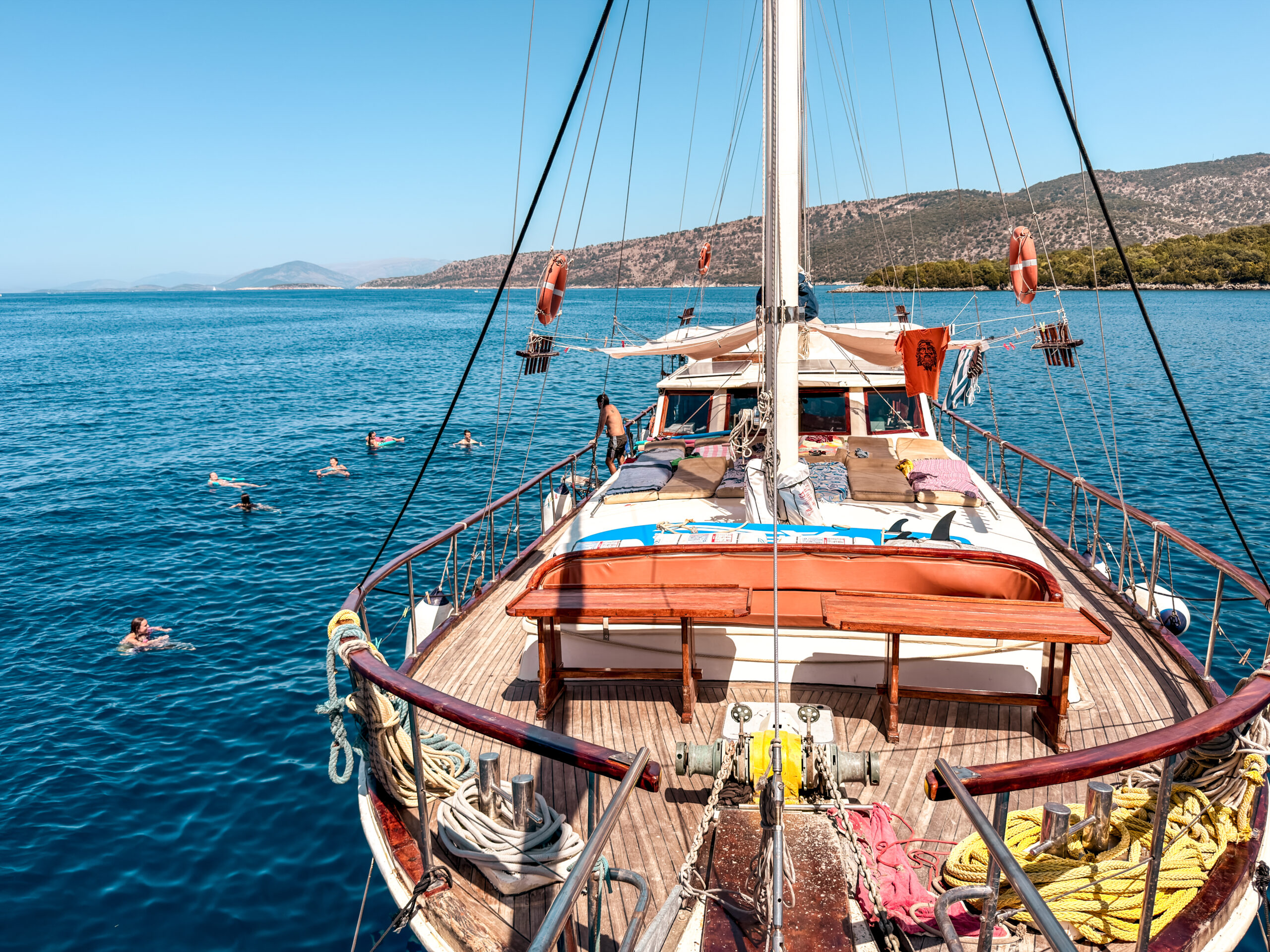 View from a sailing yacht deck watching swimmers in bright blue sea with a rocky coastal landscape in the distance.