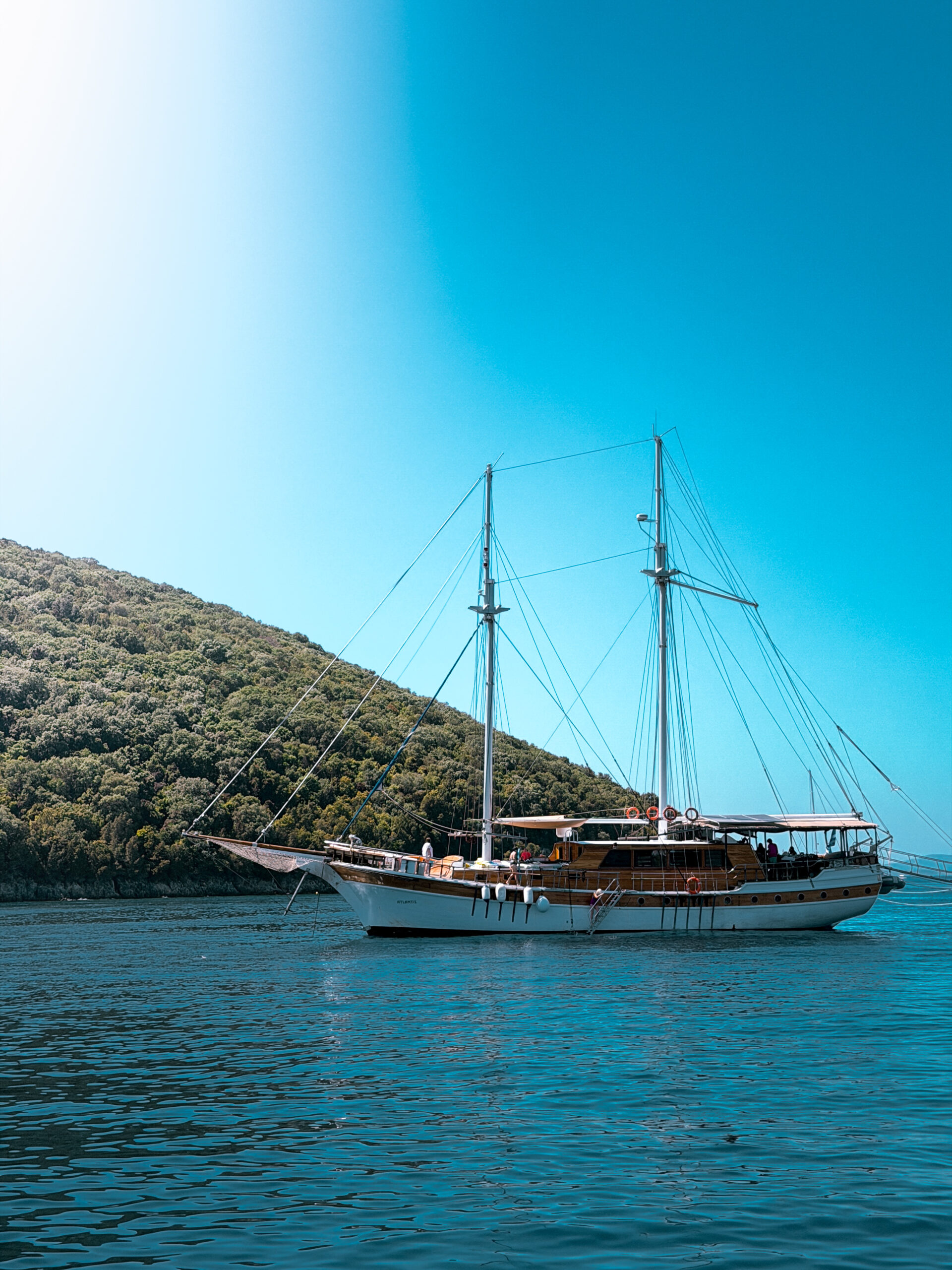 image00025 | Sail in Greece™ Sailing yacht moored in calm blue waters near a forested hillside under a clear blue sky, with masts and rigging visible.