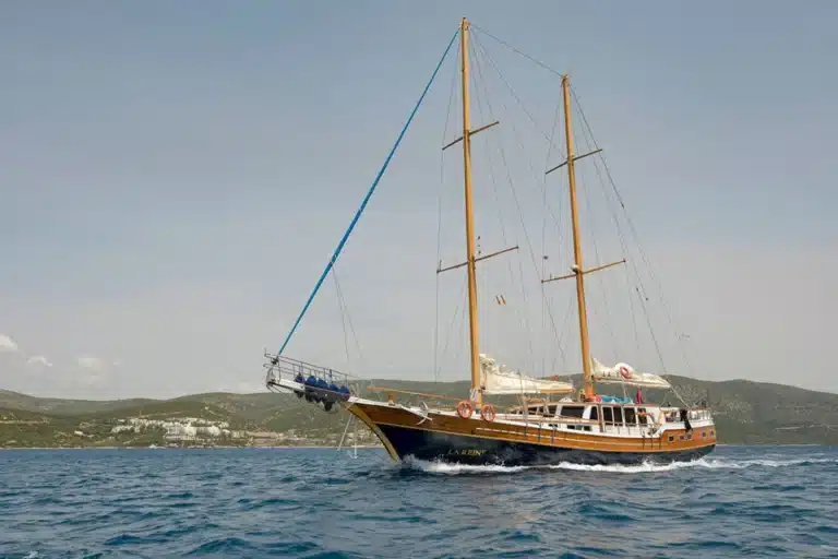 kos-left | Sail in Greece™ | Sail in Greece™ Two-masted wooden sailing yacht gliding over blue water with a distant hilly coastline in the background, name La Reina on the hull.