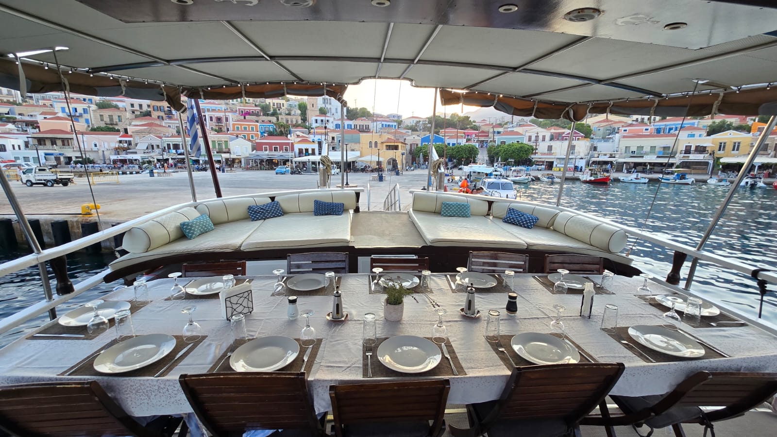 WhatsApp Image 2026-08-11 at 124529 PM 1 | Sail in Greece™ Dining table aboard a yacht with place settings and glassware, facing a curved cream sofa and a harbor with colorful waterfront buildings in the background.