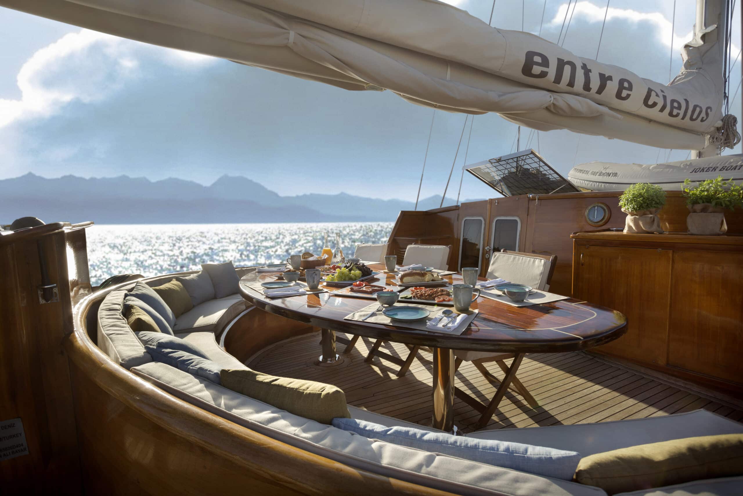 Luxurious yacht deck dining table set for a meal with plates, cups, and fruit, ocean and distant mountains in the background.